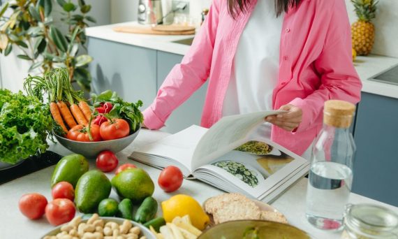 A person browsing a cookbook surrounded by fresh vegetables in a bright kitchen setting.